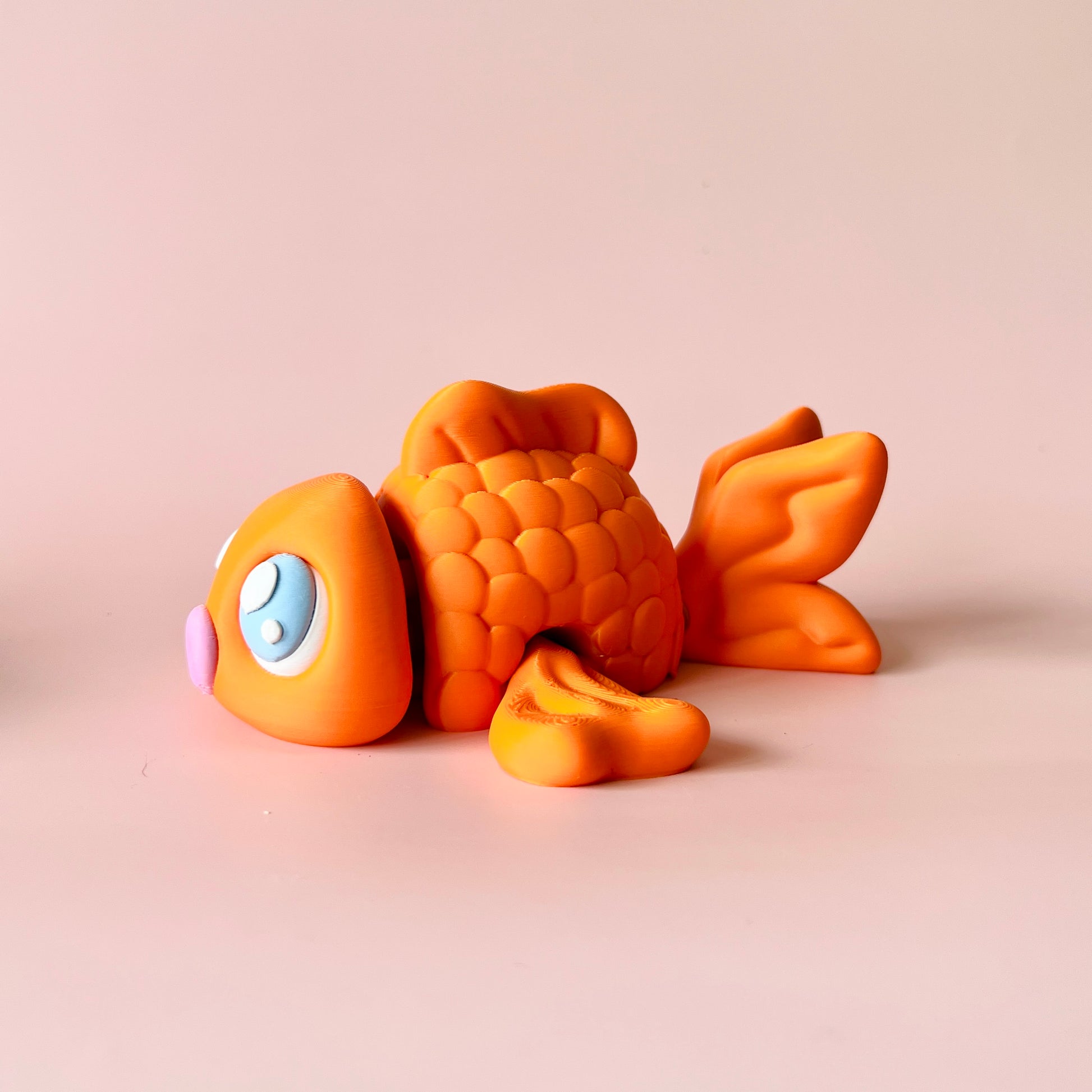 Vibrant orange fish with large blue eyes and a pink nose, resting on a soft pastel background.