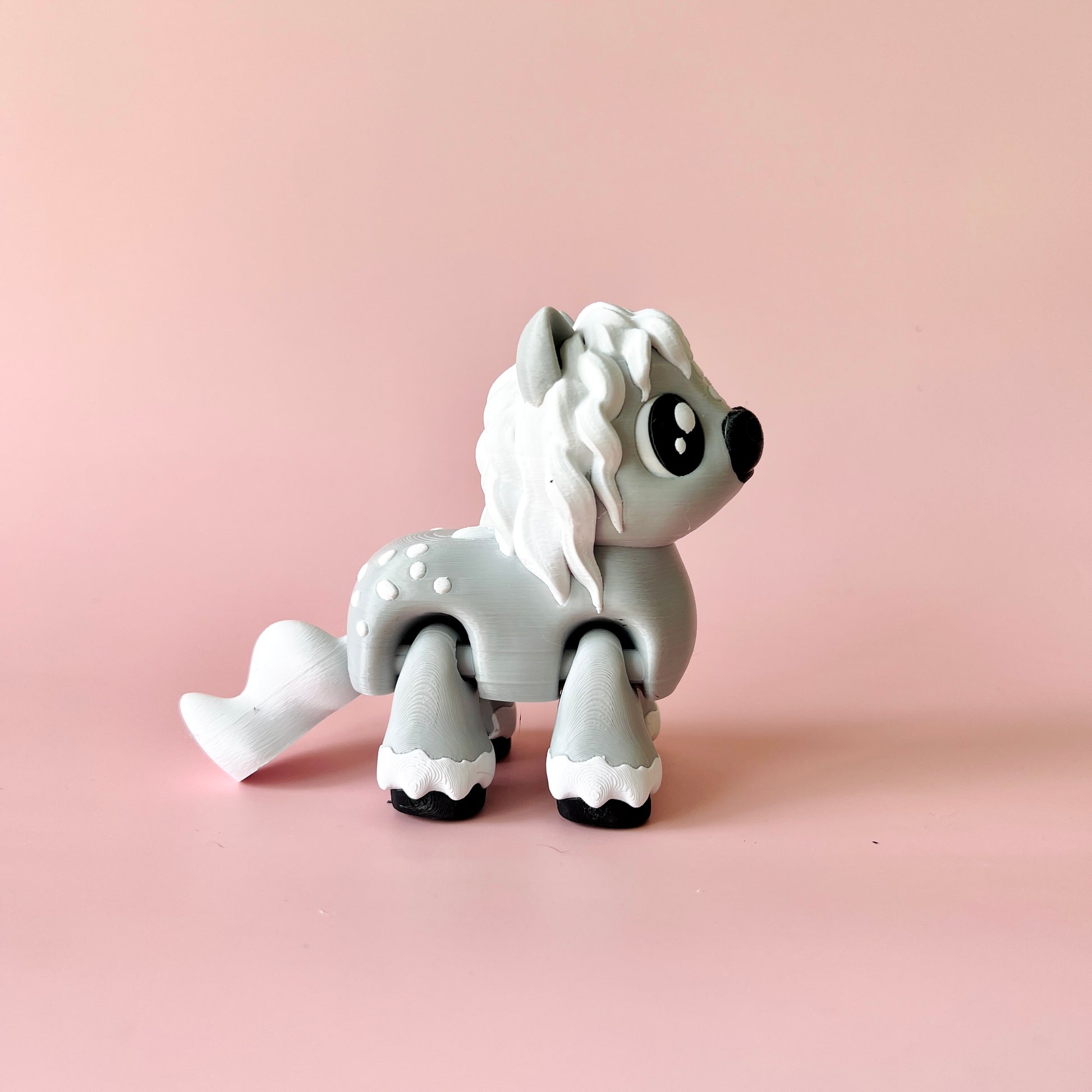 A gray, stylized pony figure with a flowing mane stands against a soft pink background, showcasing playful details.