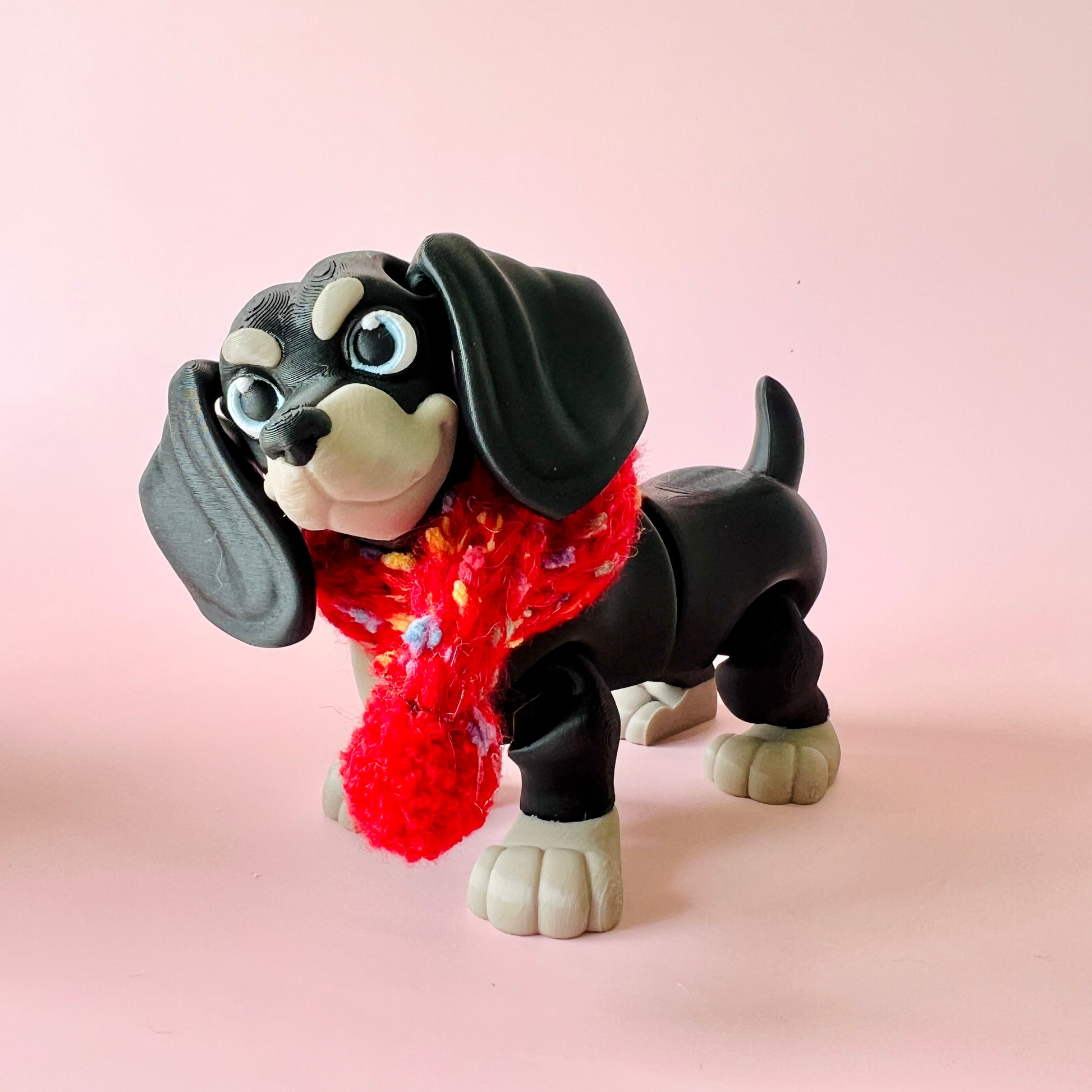 A playful black dog figure wearing a colorful knitted scarf against a soft pink background.