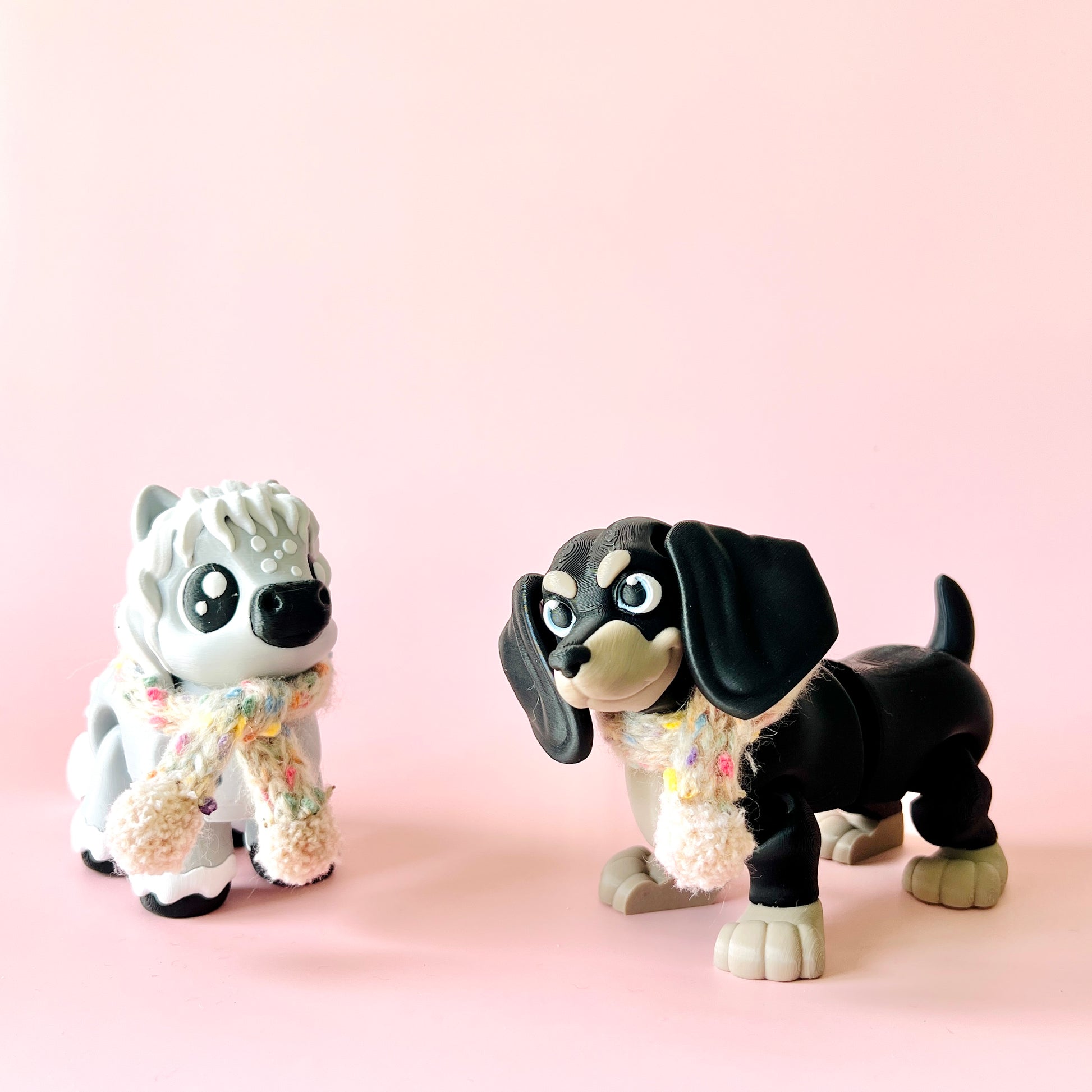 Two whimsical dog figurines wearing colorful scarves stand against a soft pink background, exuding playful charm.