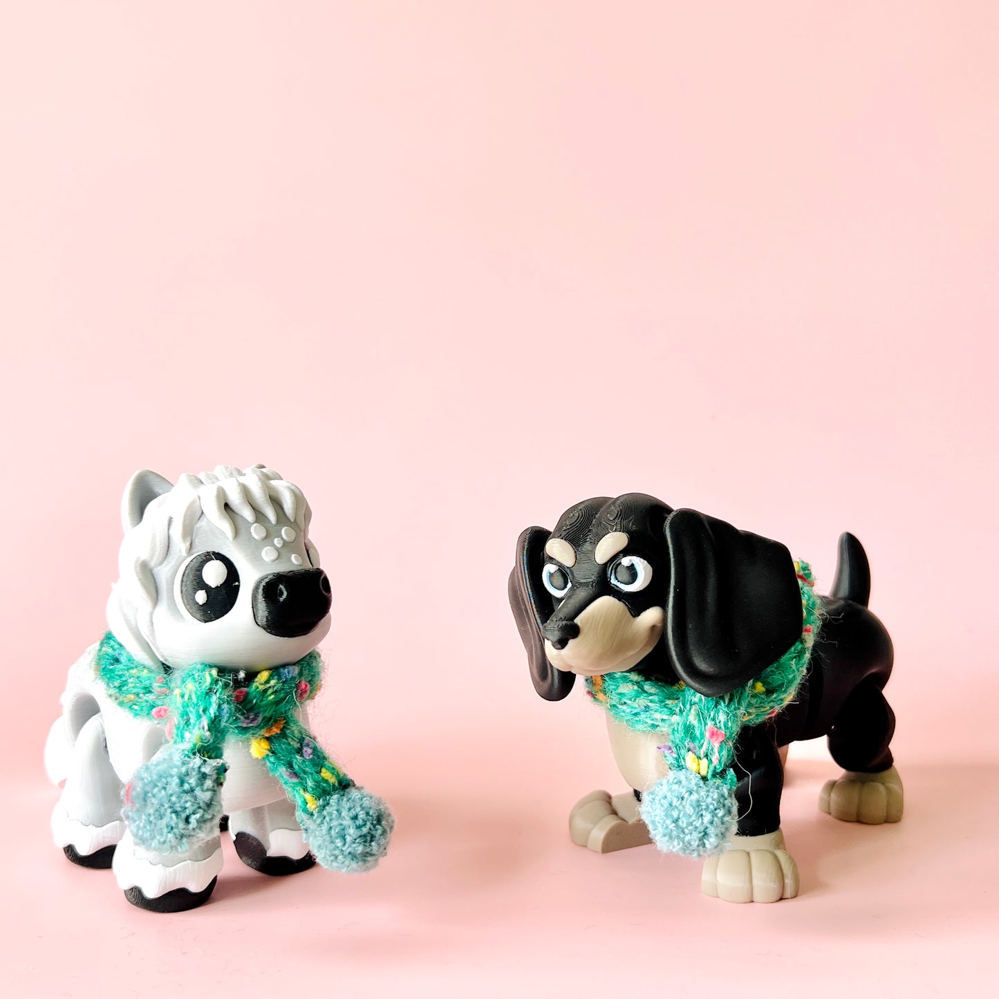 Two playful dog figurines, one white with a fluffy mane and one black, both wearing colorful knitted scarves.