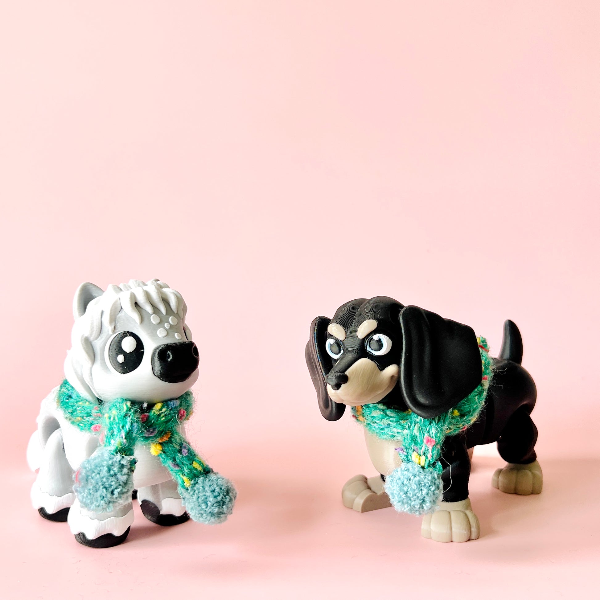 Two playful dog figurines, one white with a fluffy mane and one black, both wearing colorful knitted scarves.