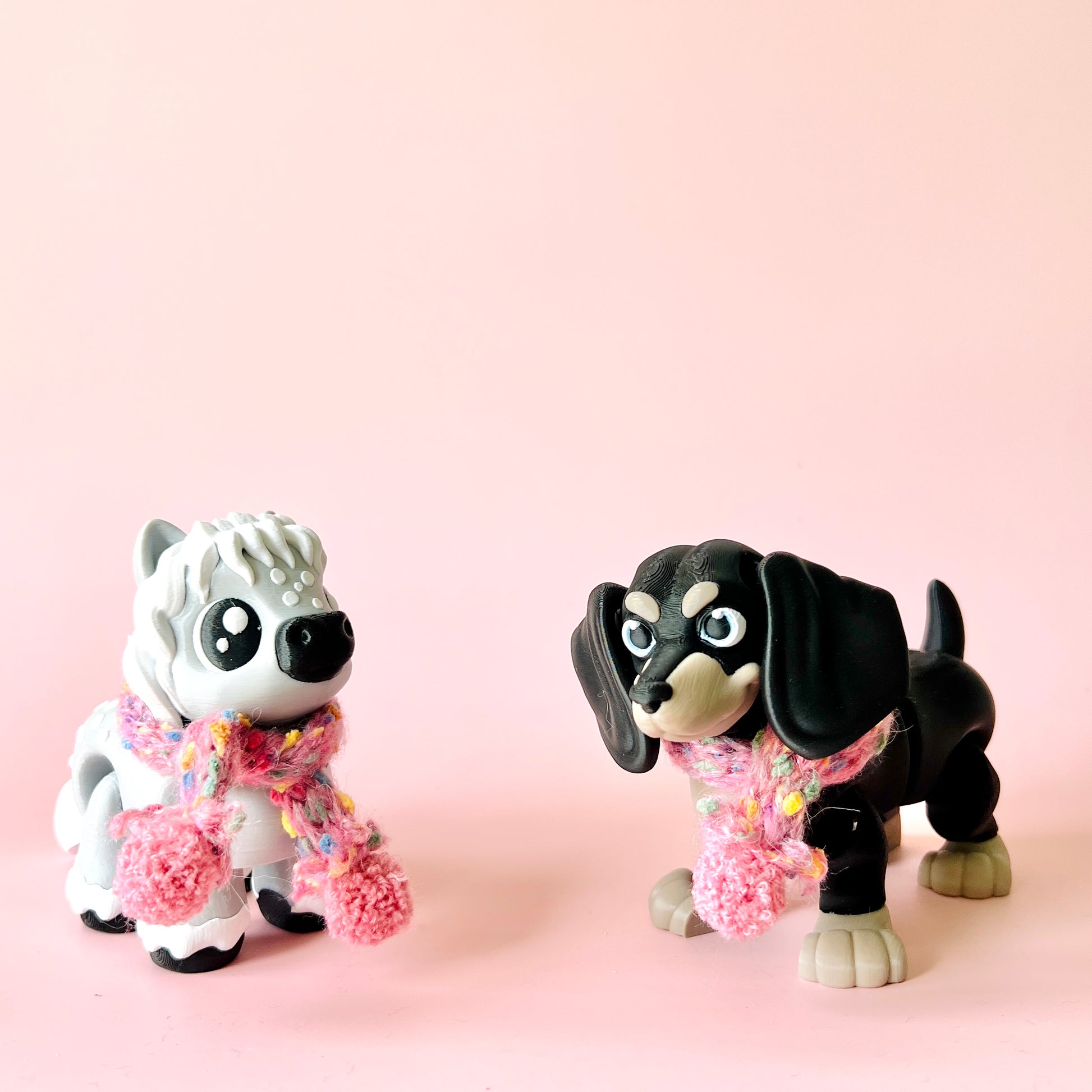 Two playful figurines resembling dogs, wearing colorful scarves, set against a soft pink background.