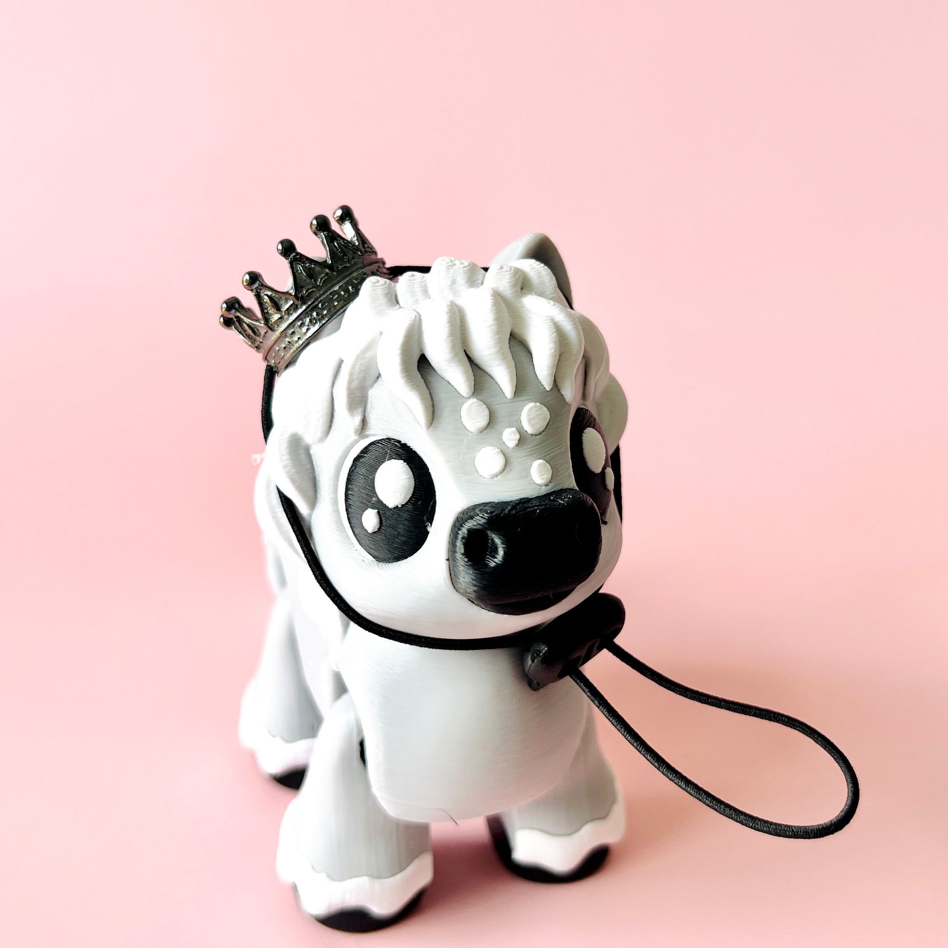 A whimsical white creature with a crown, black accents, and a leash, set against a soft pink background.