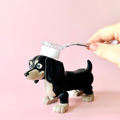 A playful black and tan dog figurine with a toothbrush held above its head, set against a soft pink background.