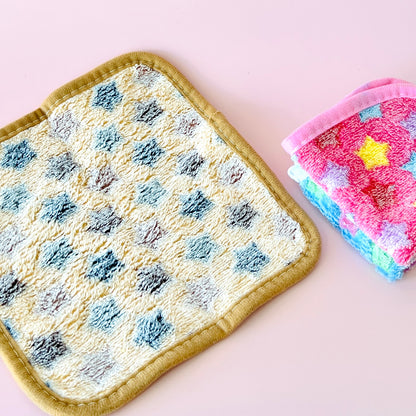 Colorful cloths with star patterns, one folded in pink and blue, next to a textured yellow and blue square on a pale backgrou