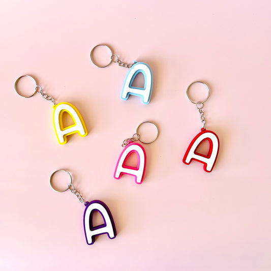 Colorful letter "A" keychains in various hues arranged on a soft pink background.
