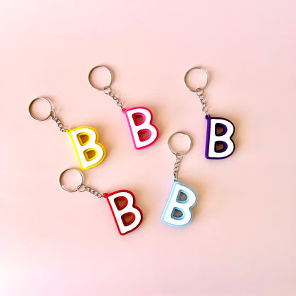Five colorful keychains shaped like the letter "B" arranged on a soft pink background.