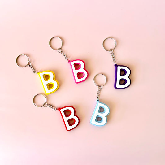 Five colorful keychains shaped like the letter "B" arranged on a soft pink background.