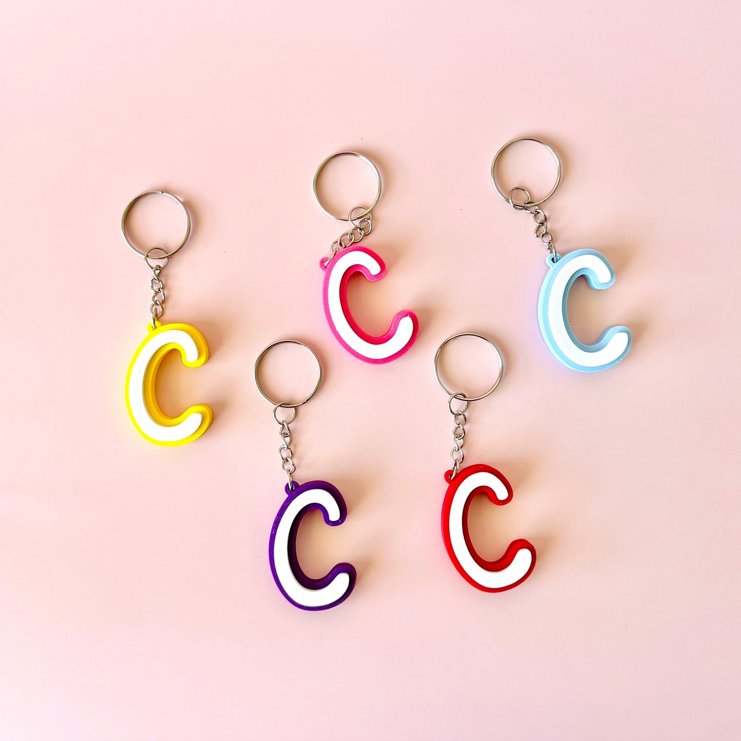 Five colorful letter "C" keychains in yellow, pink, purple, red, and blue against a soft pink background.