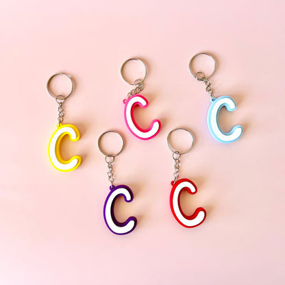 Five colorful letter "C" keychains in yellow, pink, purple, red, and blue against a soft pink background.