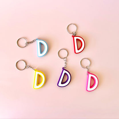 Five colorful keychains shaped like the letter "D" on a soft pink background.