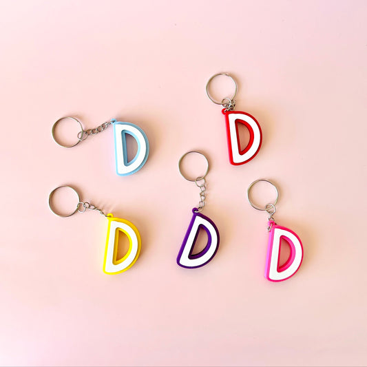 Five colorful keychains shaped like the letter "D" on a soft pink background.
