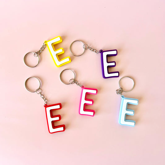 Colorful letter "E" keychains in yellow, red, pink, purple, and blue against a soft pink background.
