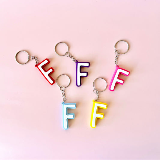 Colorful letter "F" keychains in red, purple, pink, blue, and yellow on a soft pink background.
