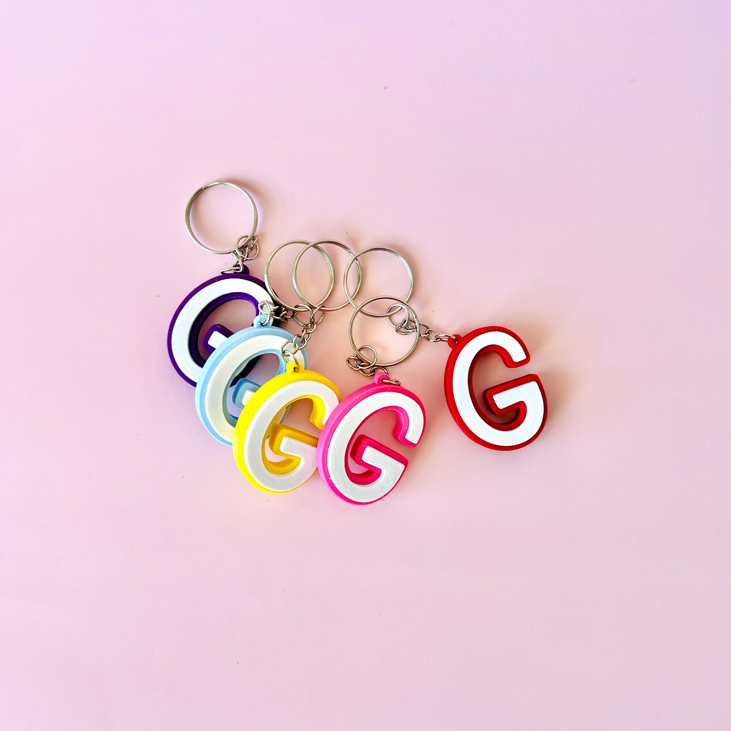 Colorful letter 'G' keychains in purple, blue, yellow, pink, and red on a soft pink background.