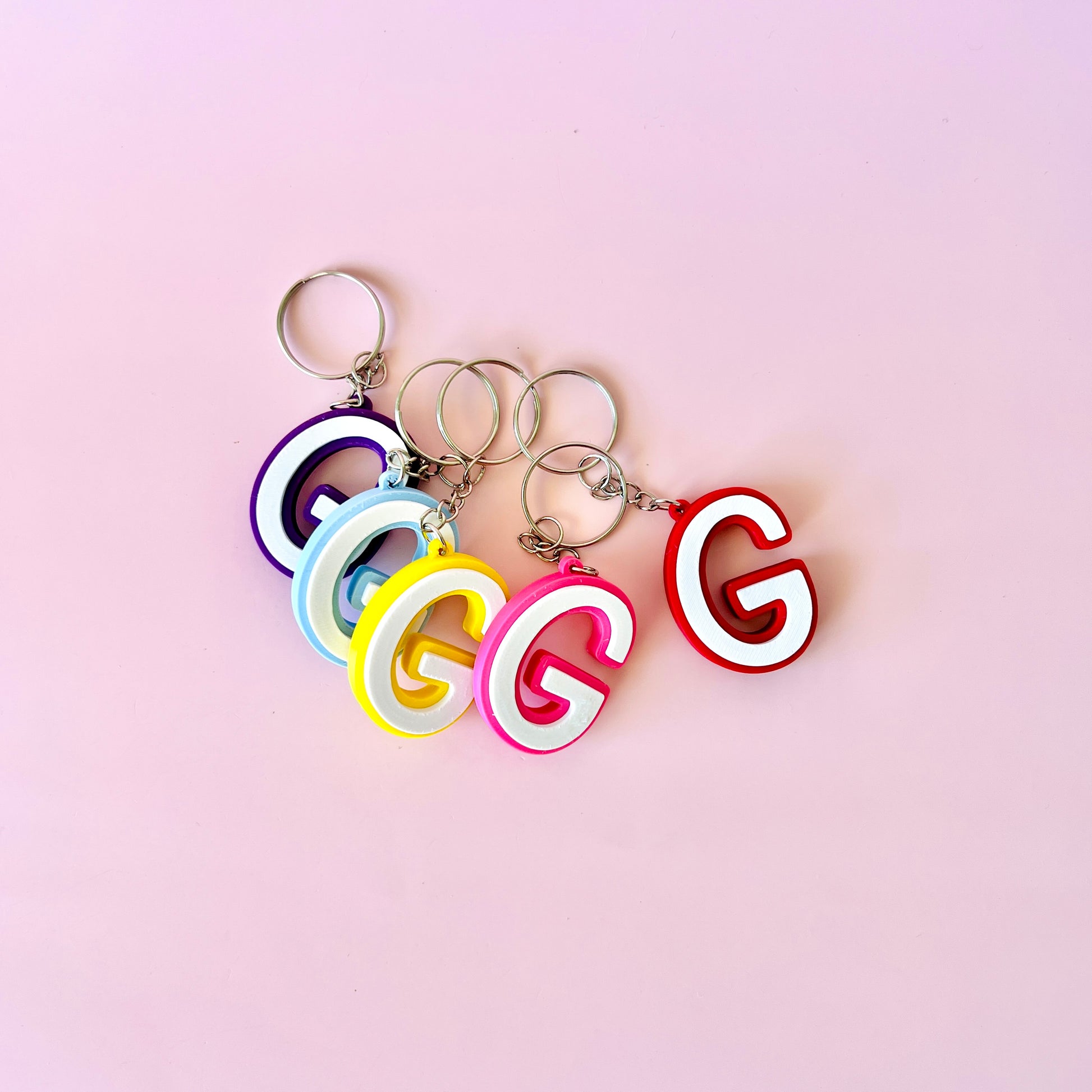 Colorful letter 'G' keychains in purple, blue, yellow, pink, and red on a soft pink background.