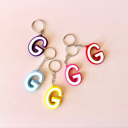 Colorful letter "G" keychains in purple, blue, red, pink, and yellow on a soft pink background.