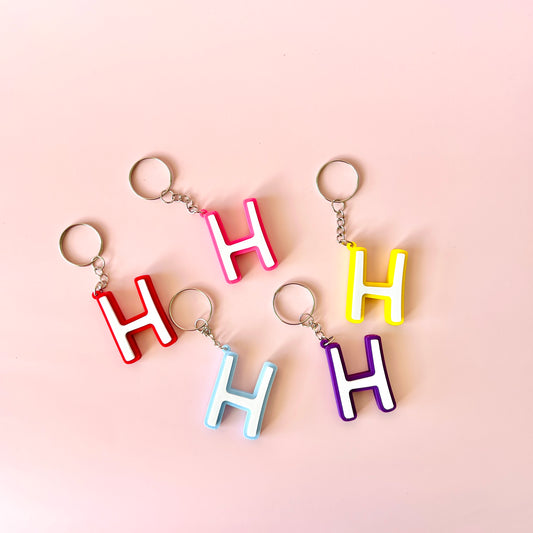 Colorful letter "H" keychains in red, pink, yellow, purple, and blue arranged on a soft pink background.