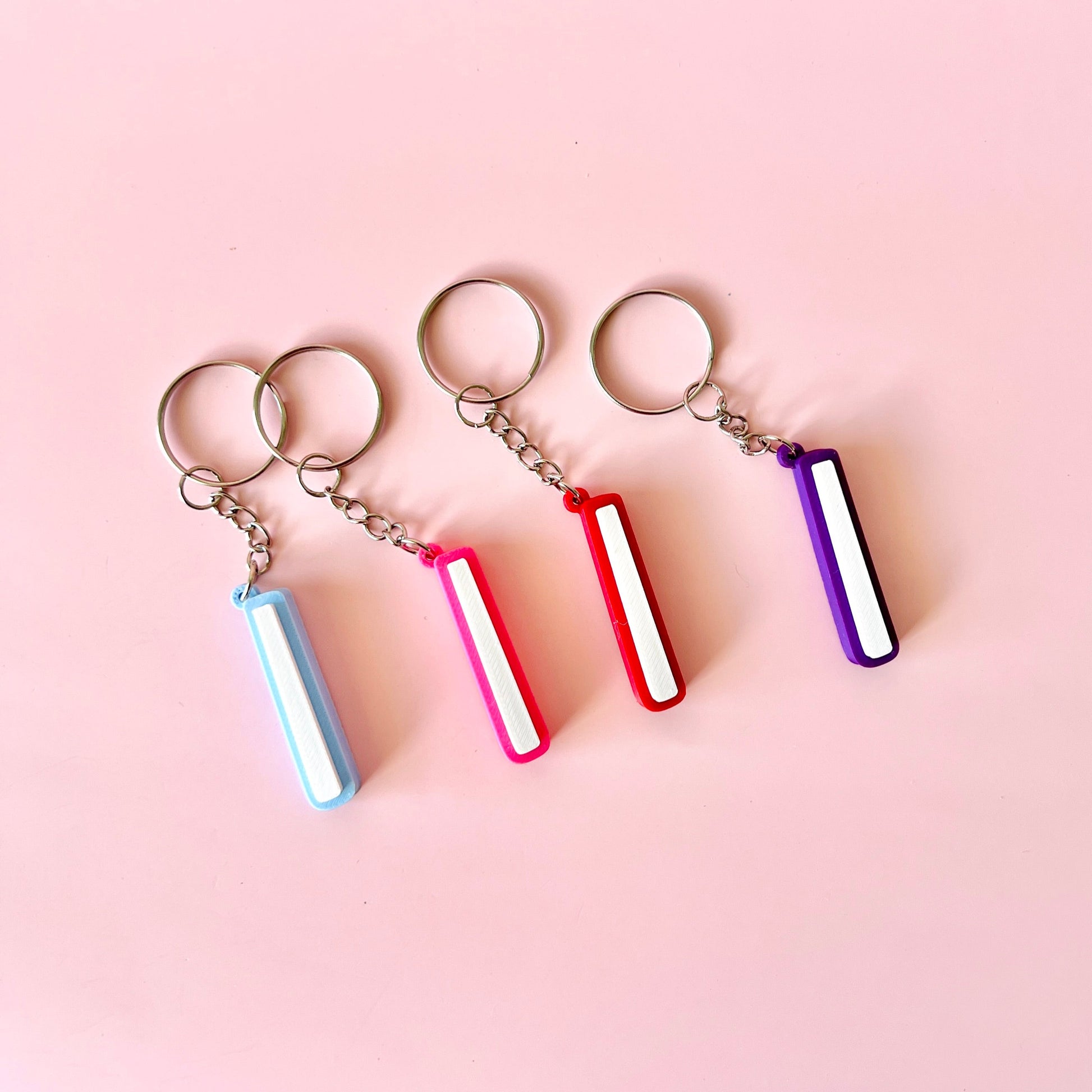 Four vibrant keychains in blue, pink, red, and purple hang against a soft pink background, showcasing playful charm.