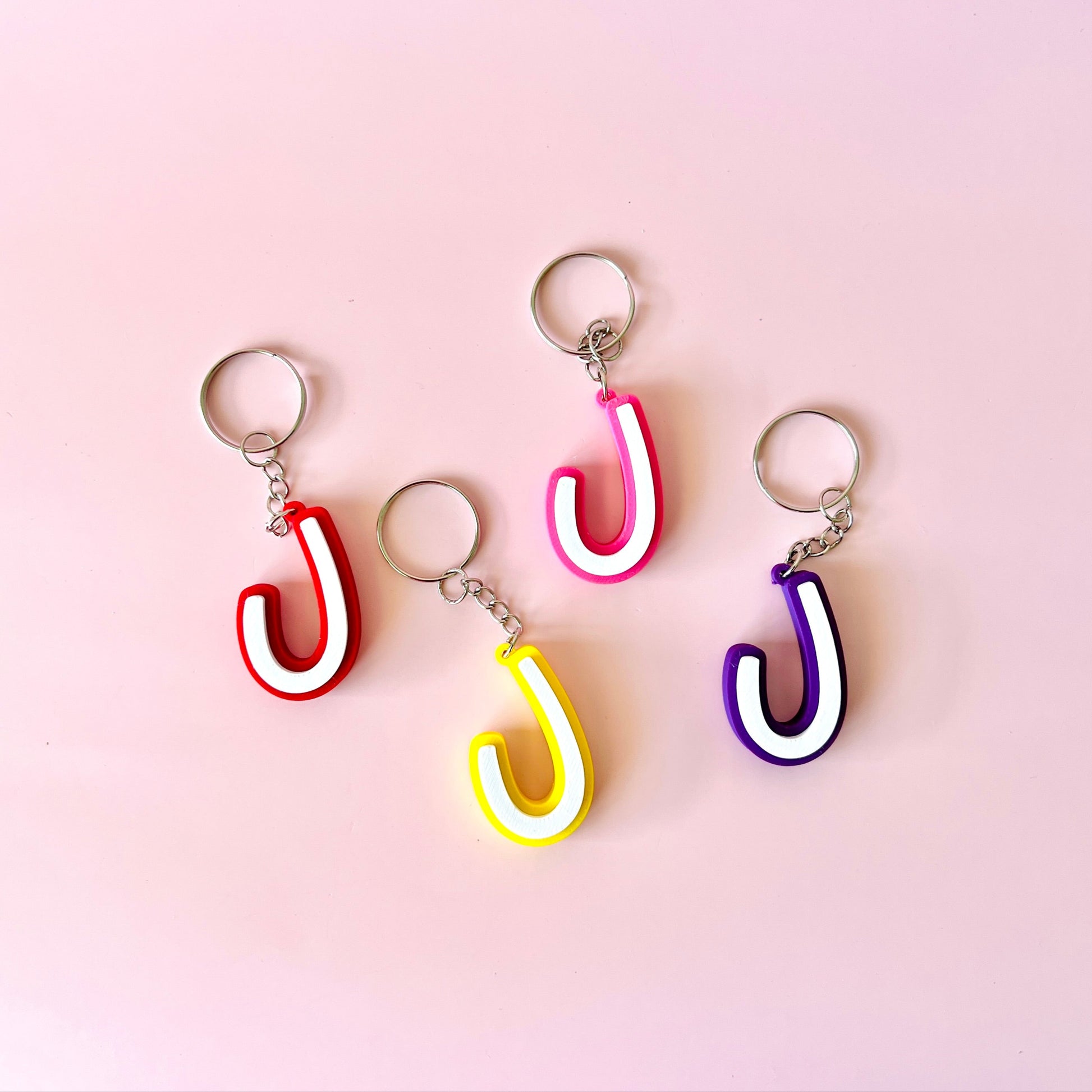 Four colorful keychains shaped like the letter "J" on a soft pink background.
