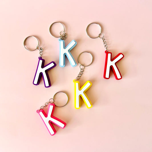 Colorful keychains shaped like the letter "K" in various hues on a soft pink background.
