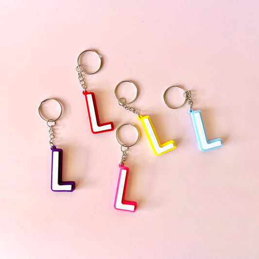 Five colorful keychains featuring the letter "L" in various shades on a soft pink background.