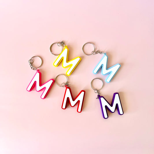 Colorful letter "M" charms in pink, yellow, blue, red, and purple arranged on a soft pink background.