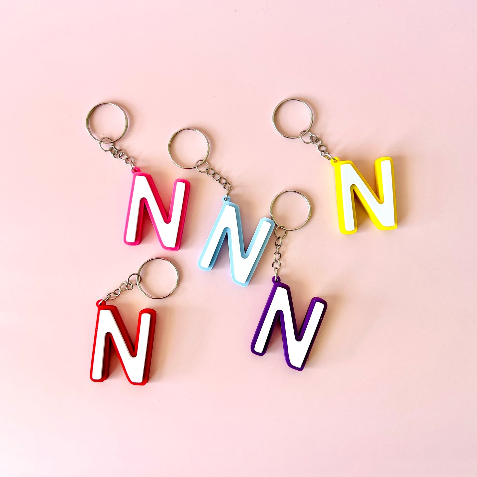 Five colorful keychains shaped like the letter "N" displayed on a soft pink background.