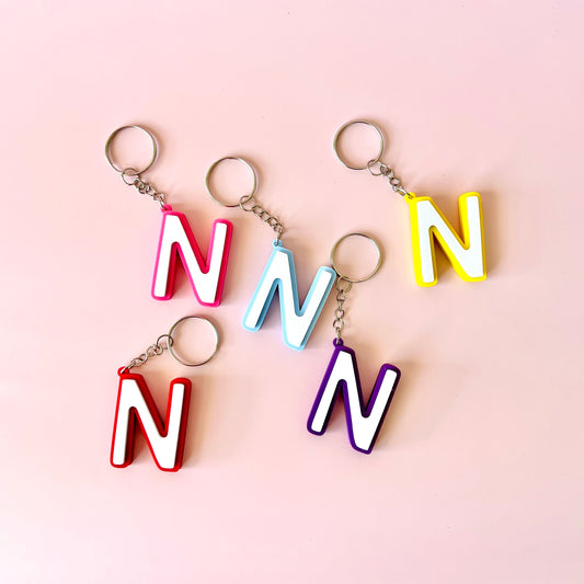 Five colorful keychains shaped like the letter "N" displayed on a soft pink background.