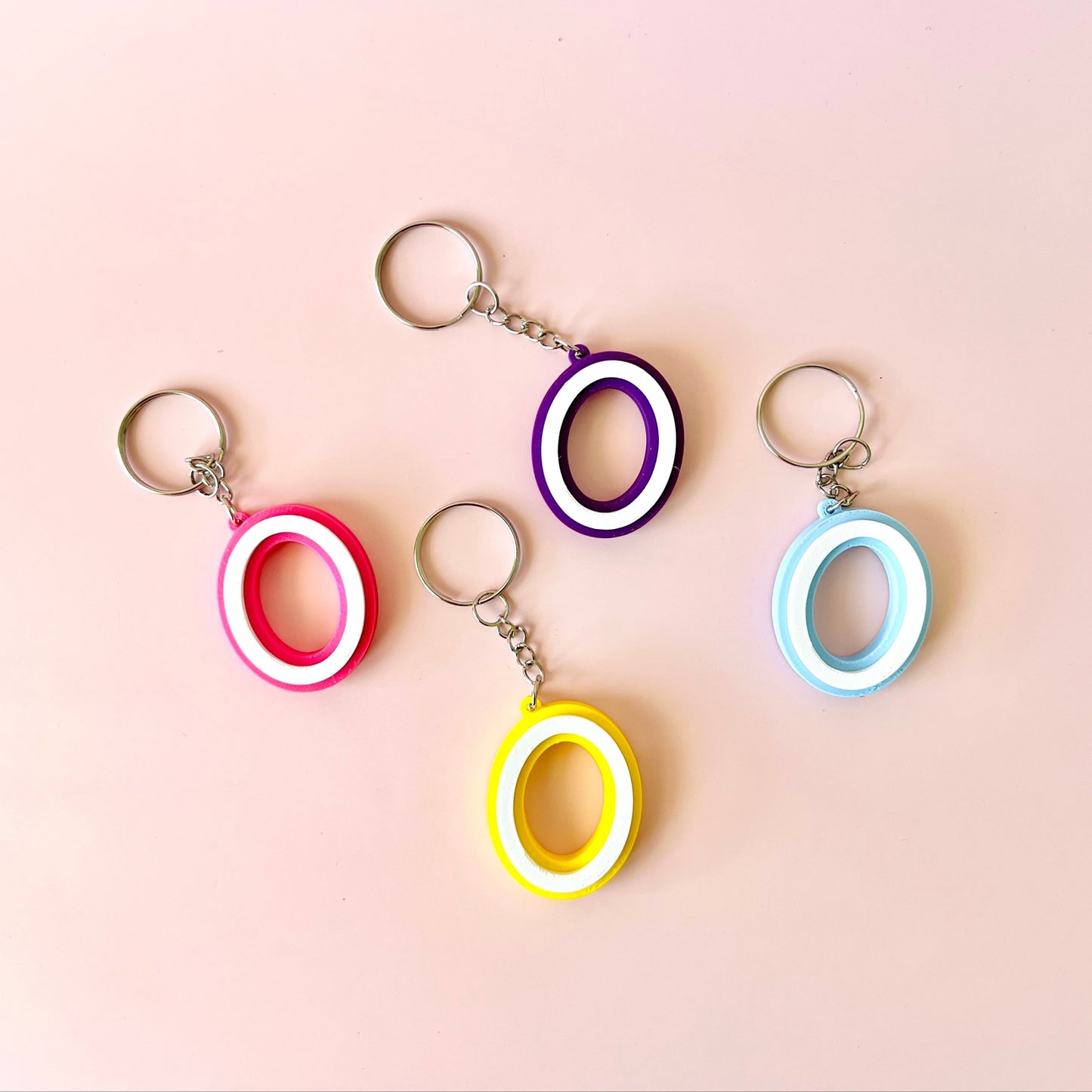 Four colorful keychains with circular shapes in pink, purple, yellow, and blue against a soft pink background.