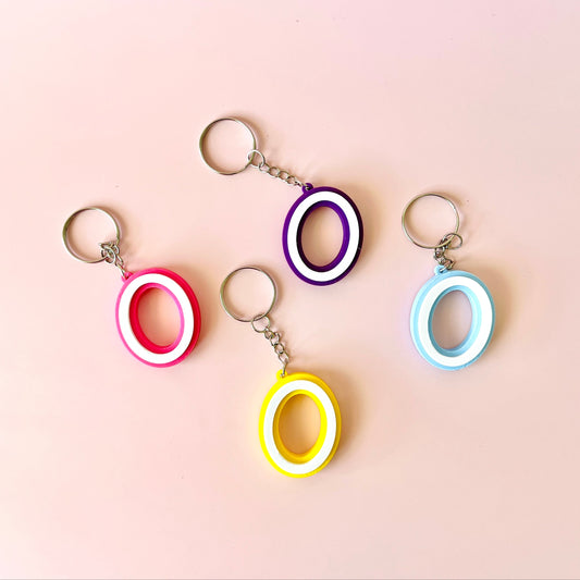 Four colorful keychains with circular shapes in pink, purple, yellow, and blue against a soft pink background.