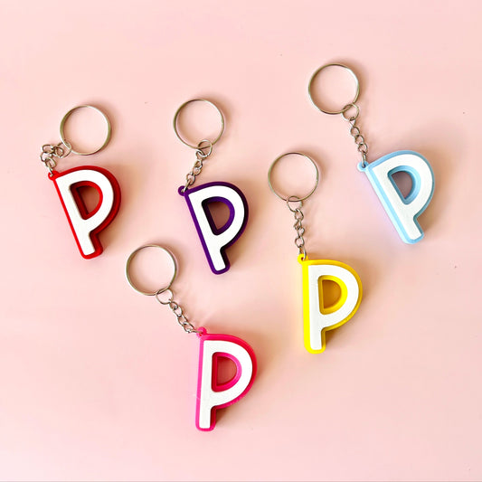 Five colorful letter "P" keychains in red, purple, pink, yellow, and blue against a soft pink background.