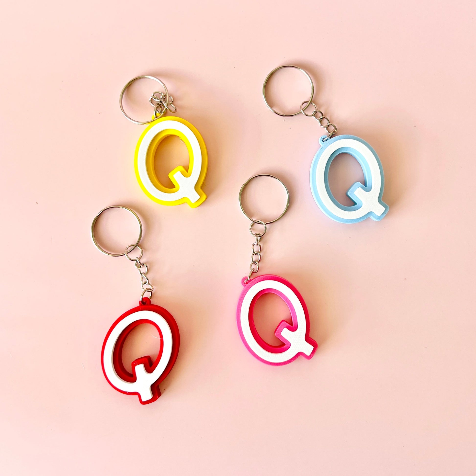 Four colorful keychains shaped like the letter "Q" on a soft pink background.