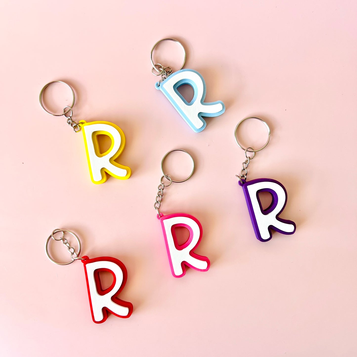 Five colorful keychains shaped like the letter "R" arranged on a soft pink background.