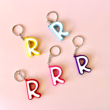 Five colorful keychains shaped like the letter "R" arranged on a soft pink background.