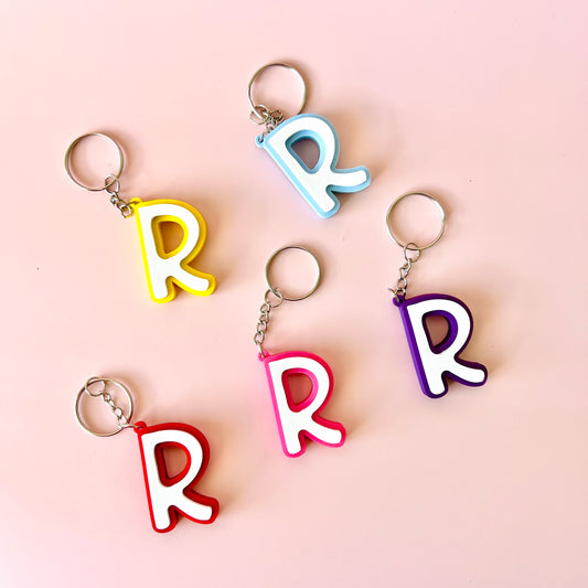Five colorful keychains shaped like the letter "R" arranged on a soft pink background.
