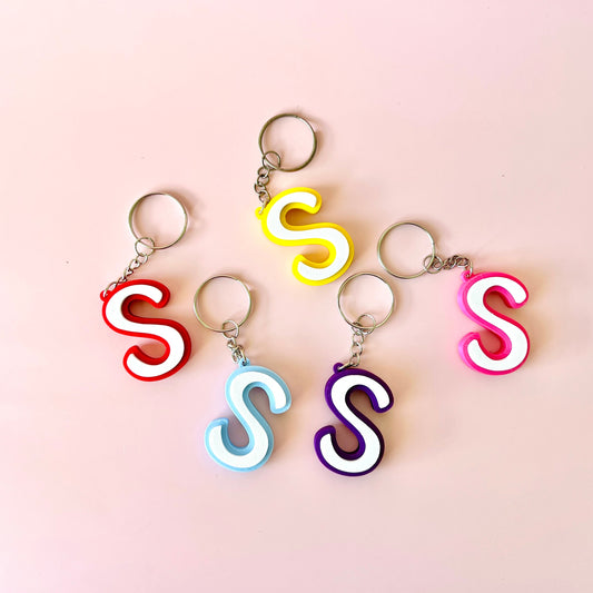 Colorful keychains shaped like the letter "S" in various hues on a soft pink background.