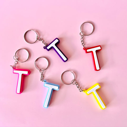 Colorful letter "T" keychains in pink, purple, red, blue, and yellow against a soft pink background.