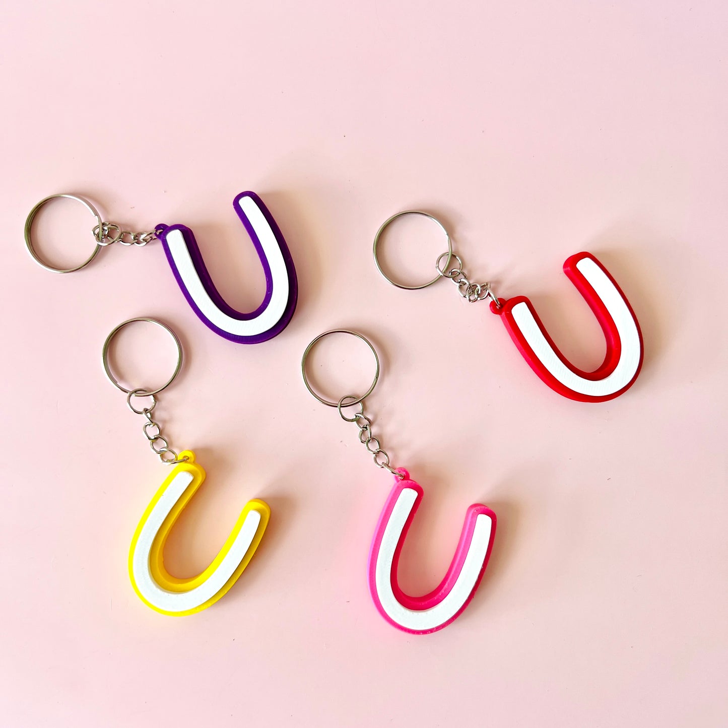 Colorful keychains shaped like the letter "U" in purple, red, pink, and yellow against a soft pink background.