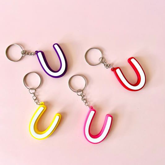 Colorful keychains shaped like the letter "U" in purple, red, pink, and yellow against a soft pink background.