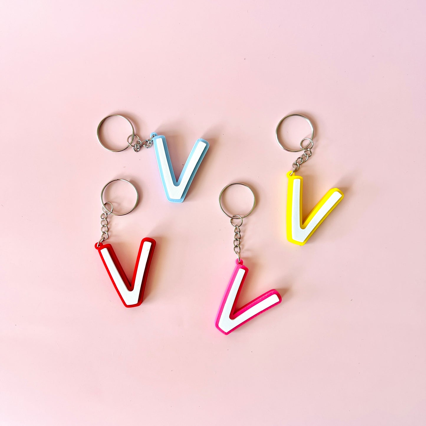 Colorful letter "V" keychains in red, pink, blue, and yellow on a soft pink background.