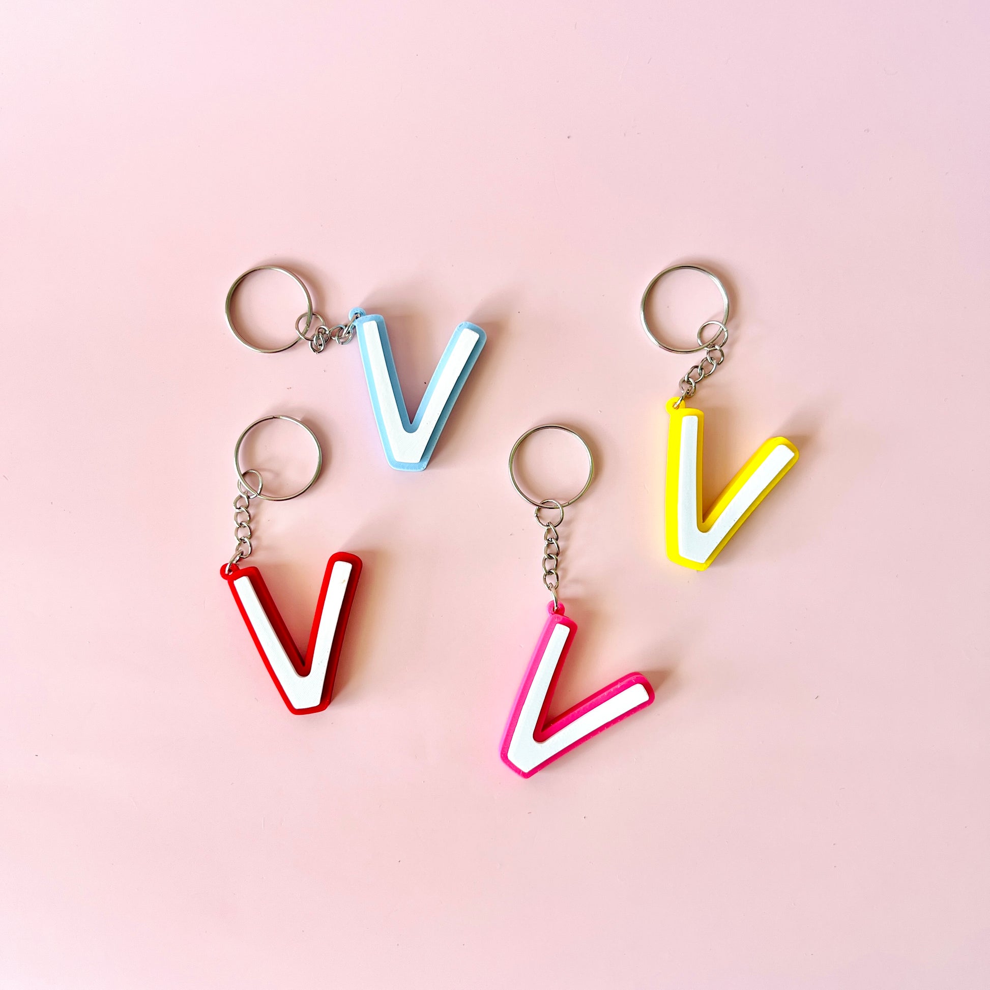 Colorful letter "V" keychains in red, pink, blue, and yellow on a soft pink background.