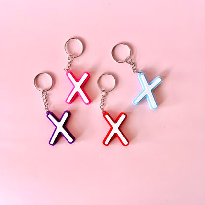 Colorful keychains in pink, purple, red, and blue shaped like the letter "X" on a soft pink background.