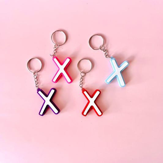 Colorful keychains in pink, purple, red, and blue shaped like the letter "X" on a soft pink background.