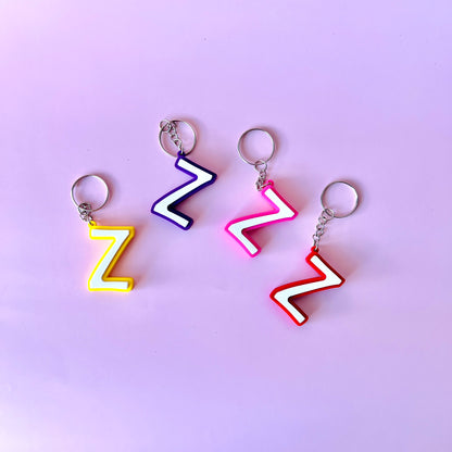 Four colorful, zigzag-shaped keychains in yellow, purple, pink, and red against a soft pink background.
