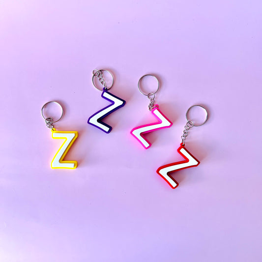 Four colorful, zigzag-shaped keychains in yellow, purple, pink, and red against a soft pink background.