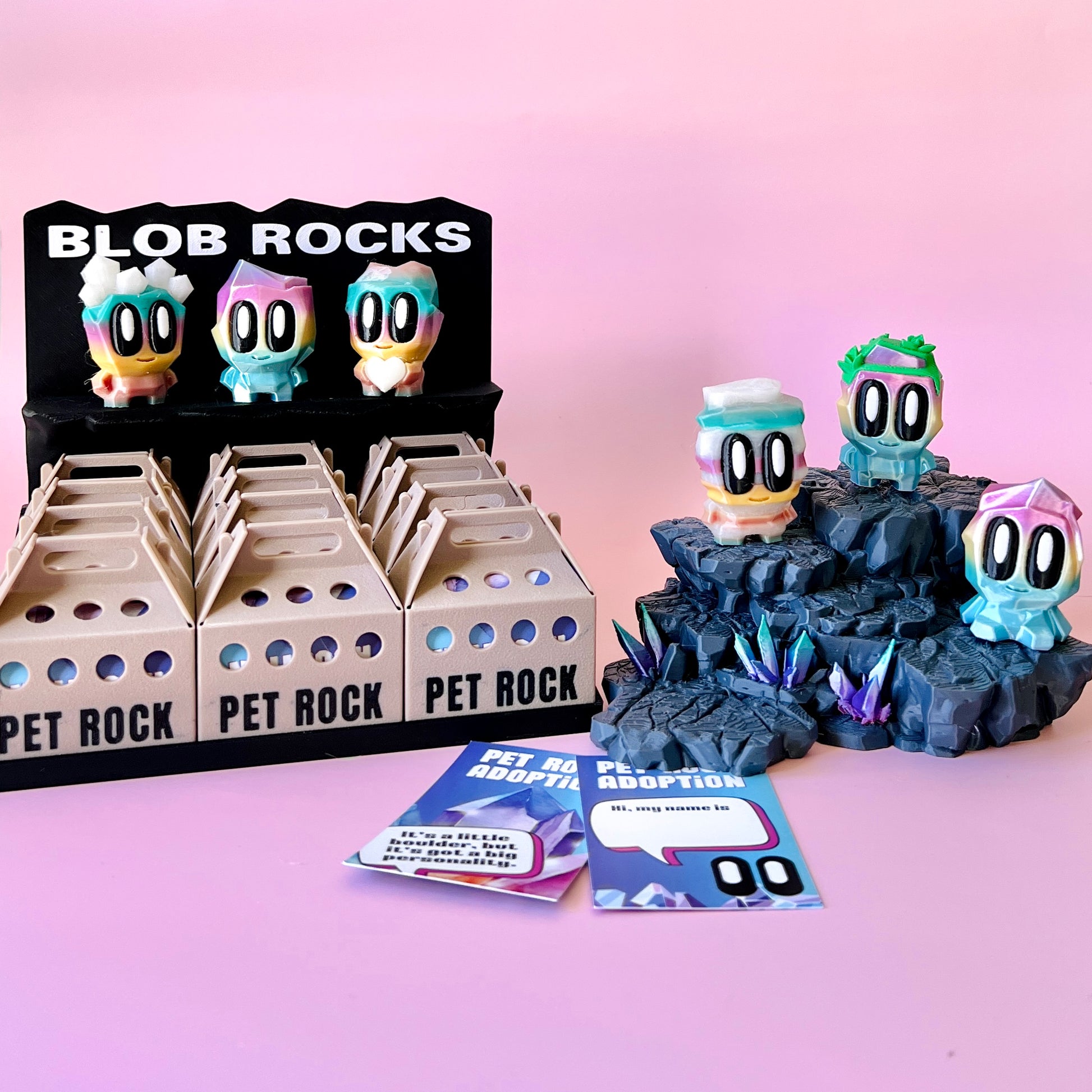 Colorful figurines displayed on a rocky base, alongside adoption cards and a playful sign on a pink background.