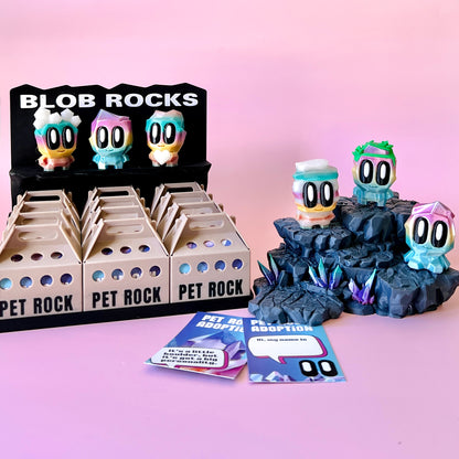 Colorful figurines displayed on a rocky base, alongside adoption cards and a playful sign on a pink background.
