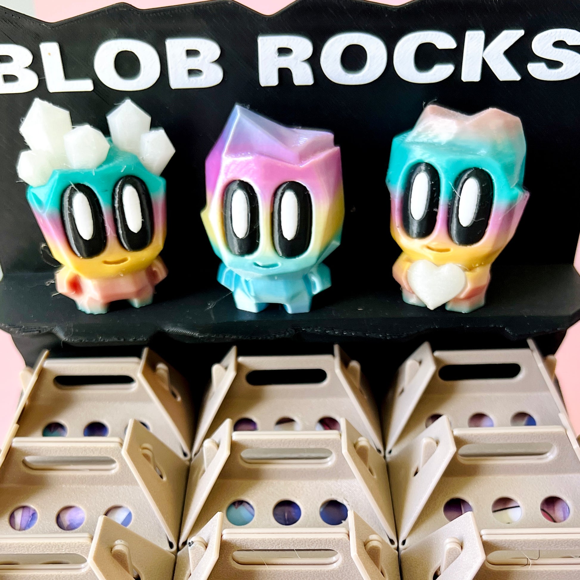 Three colorful, whimsical characters with large eyes displayed on a black backdrop, sitting atop a beige holder.